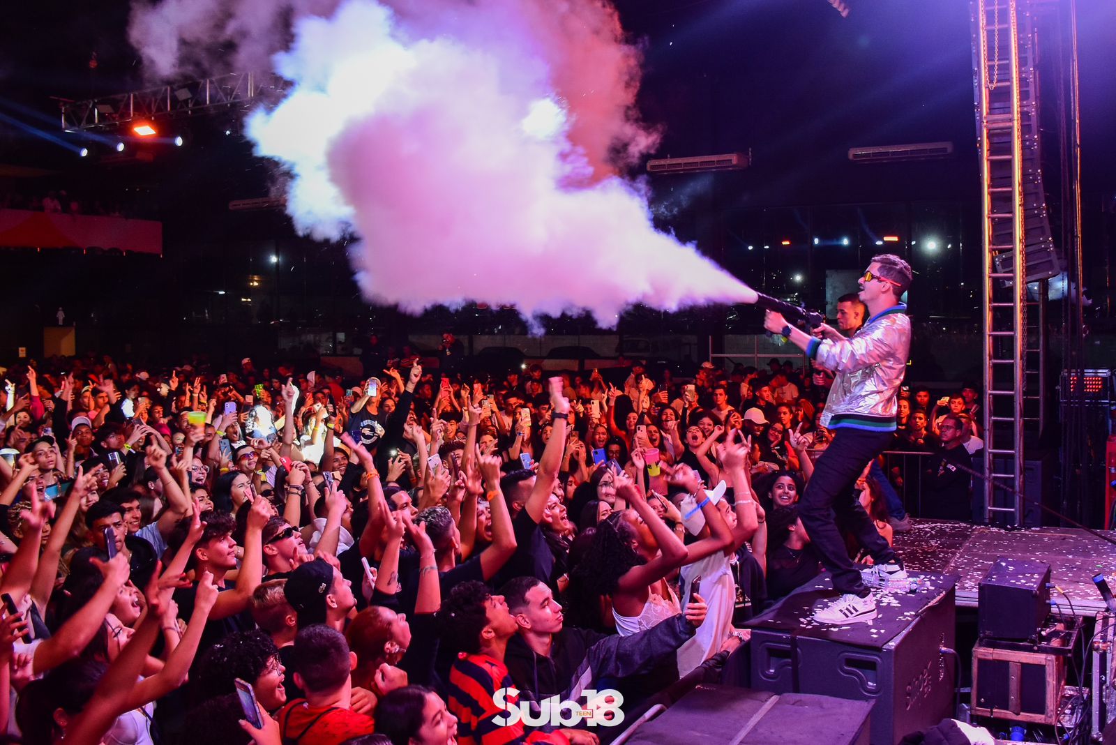 Palco da festa Sub-18 Teen, em SJC, com uma pessoa soltando uma fumaça, com jaqueta prata e o público em baixo.