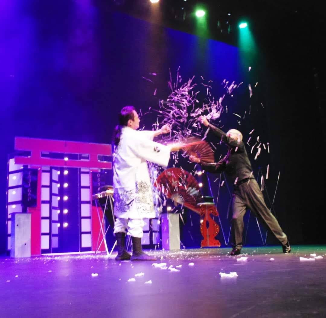 Show de mágica “The Oriental Magic Show”, atração no Teatro Colinas; foto tem fundo roxo e duas pessoas fazendo truques, com vestimentas brancas e pretas.