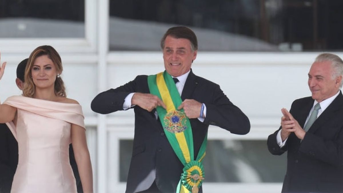 Bolsonaro com a faixa presidencial, apontando, com terno preto, ao lado de Michelle e Michel Temer.