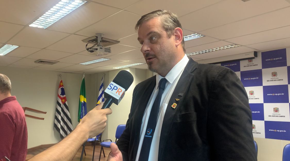 Diretor do INPE, Clésio Marcos de Nardin, com terno preto, camisa branca, gravata azul, olhando para a esquerda, falando no microfone durante entrevista sobre o evento SSP23.
