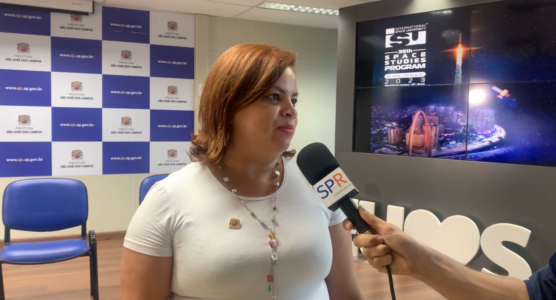 Diretora de turismo de SJC, Aline Arantes, com camisa branca, colar, olhando para a direita e uma televisão ao fundo.