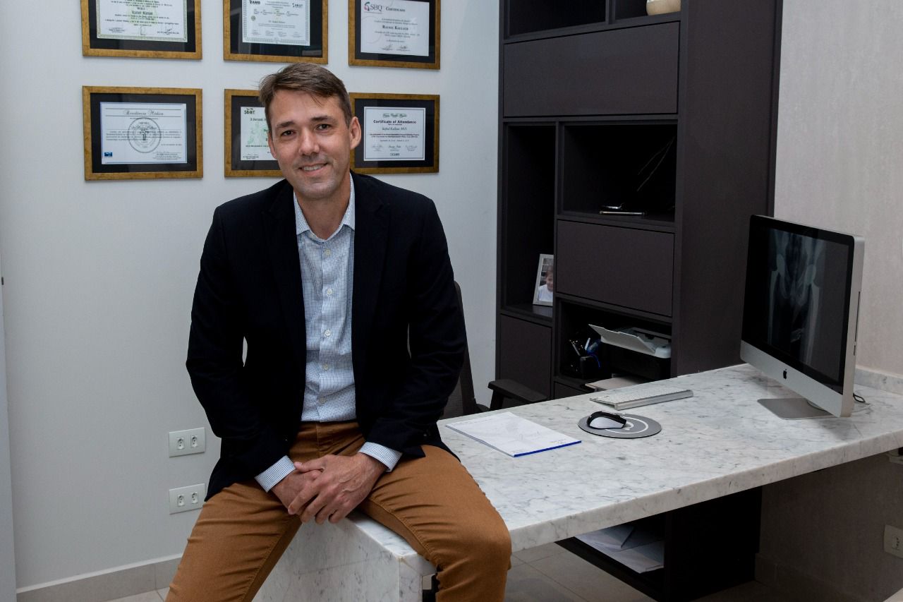 Rafael Kallaur, médico de São José dos Campos, palestrante no HIP Global Issue Network, que acontece em Portugal. Na foto ele está com calça marom, jaqueta preta, sentado em uma mesa, com quadros no fundo.