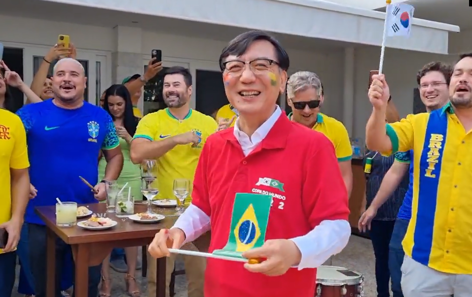 Embaixador da Coreia do Sul cantando Evidências com brasileiros, vestindo camisa vermelha, com torcedores ao fundo, vestindo amarelo.
