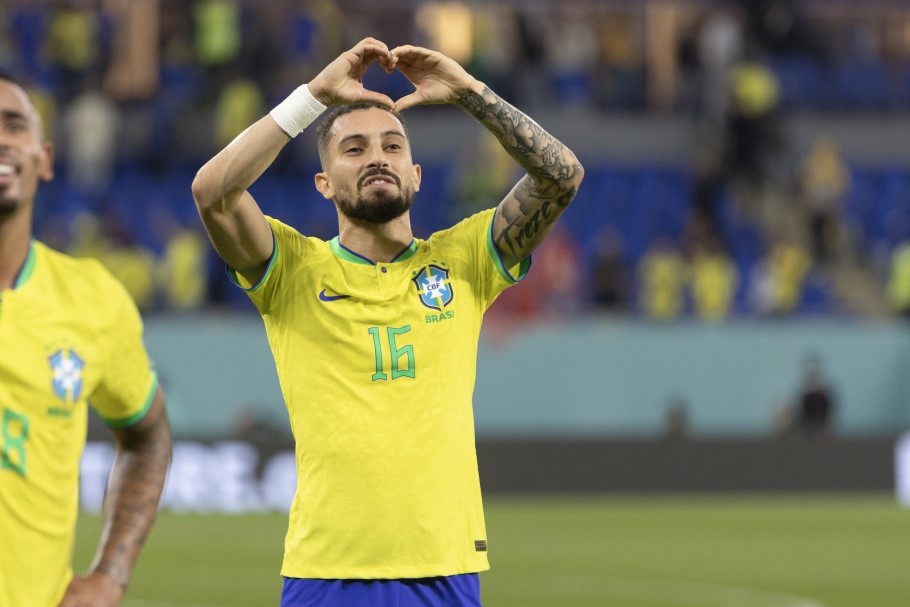 Alex Telles com a camisa amarela da Seleção Brasileira, fazendo um coração com as mãos, no gramado dentro de campo, com o fundo desfocado.