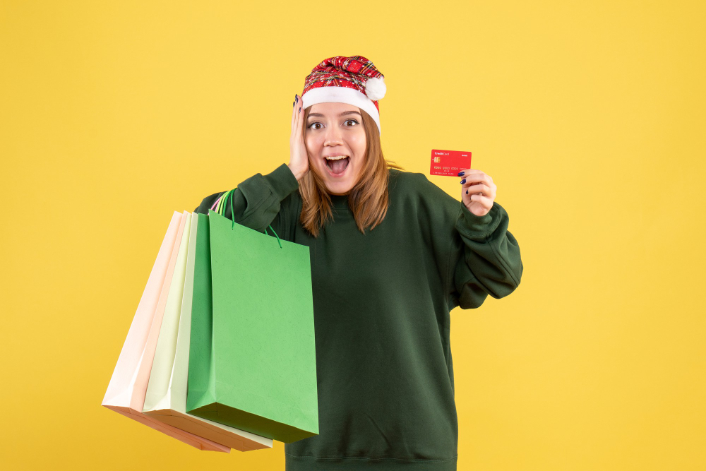 Mulher com touca de Natal, blusa verde, segurando um cartão de crédito vermelho, sacolas no outro braço e fundo amarelo; foto ilustra a matéria que diz que atendimento personalizado será decisivo para as compras de Natal.