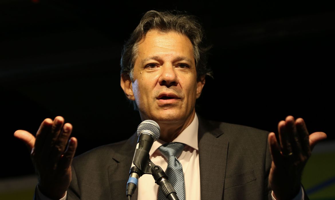Fernando Haddad com terno preto, camisa branca, gravata azul, falando no microfone, com fundo preto, olhando para a câmera; foto ilustra a matéria sobre mulheres anunciadas para o Ministério da Fazenda.