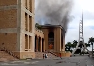 Incêndio na Basílica de Aparecida, com fumaça preta no céu, pessoas andando e o tijolo à vista à mostra.
