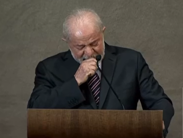 Lula chorando durante diplomação em Brasília, com tenro preto, gravata listrada, mão no queixo e microfone preto.