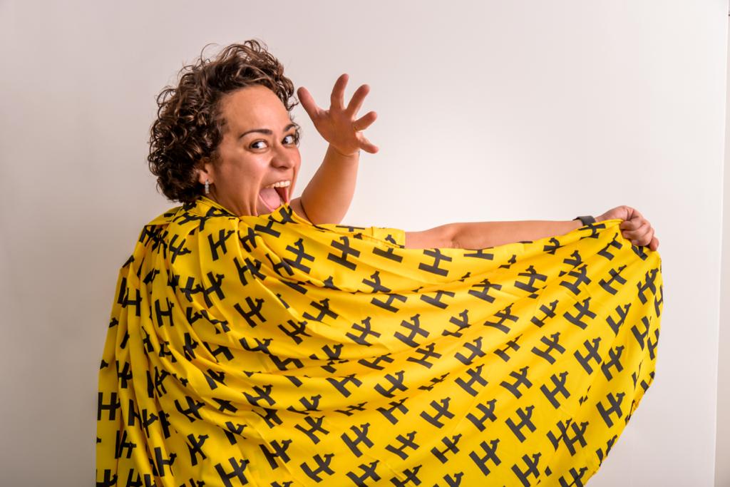 Maryana com Y com um vestido amarelo, com escritas pretas, com a boca aberta, olhando para a câmera, com a mão para cima, em frente a uma parede branca.
