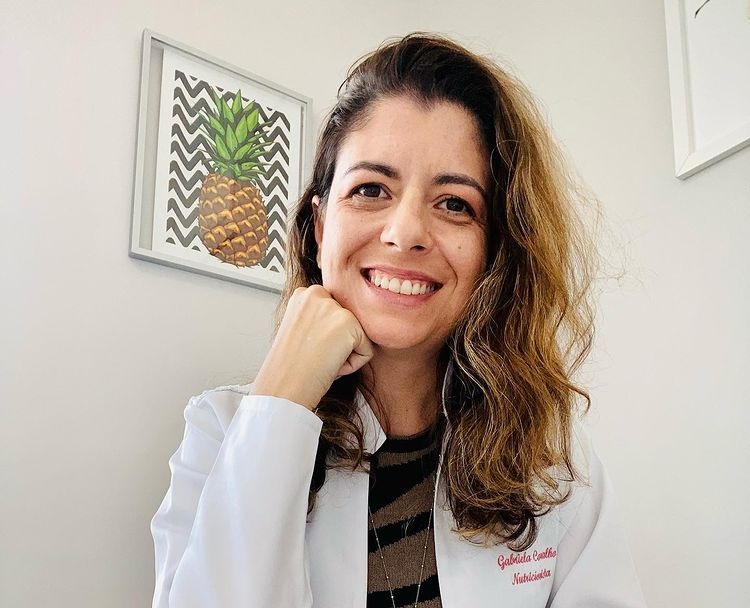 Nutricionista Gabriela Carvalho, de SJC, especialista em plant-based, com um jaleco branco, mão no queixo e fundo neutro.