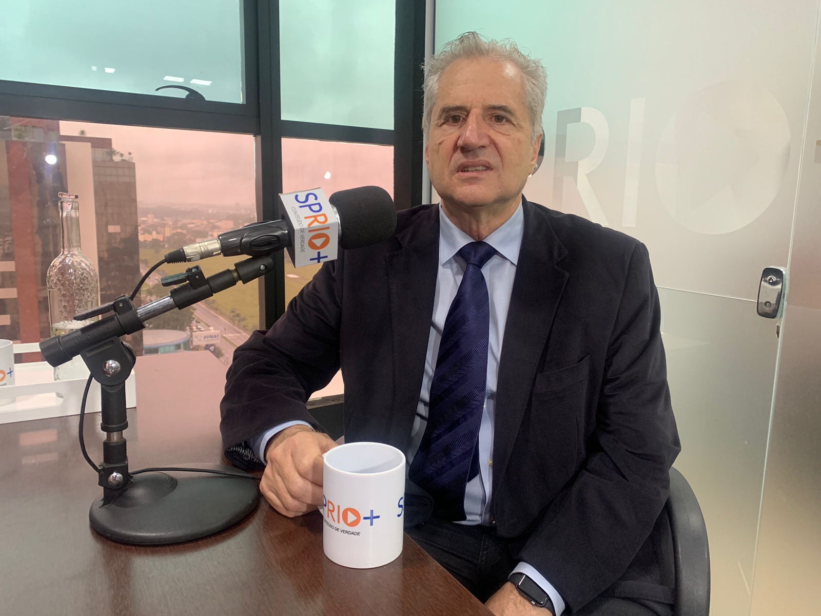 Célio Costa Vaz, CEO da Orbital Engenharia, empresa de SJC, durante entrevista, com terno preto, camisa branca, gravata azul, falando no microfone, segurando uma caneca e a cidade de São José dos Campos ao fundo.