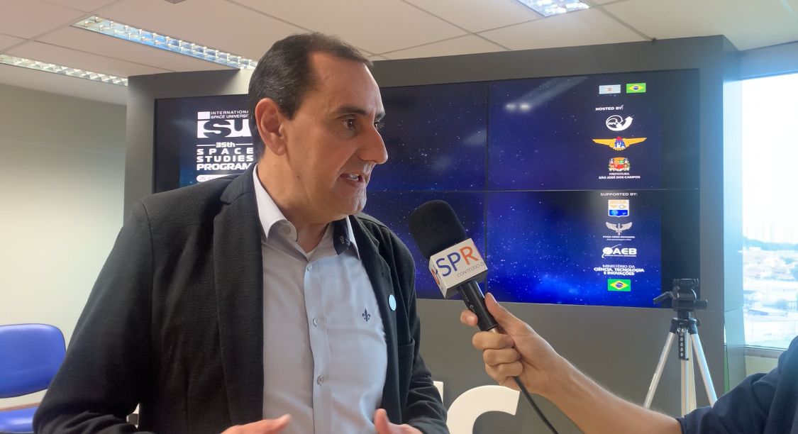 Anderson Farias, prefeito de SJC, durante entrevista sobre o evento SSP23, com terno preto, camisa azul, com um telão ao fundo e teto branco.