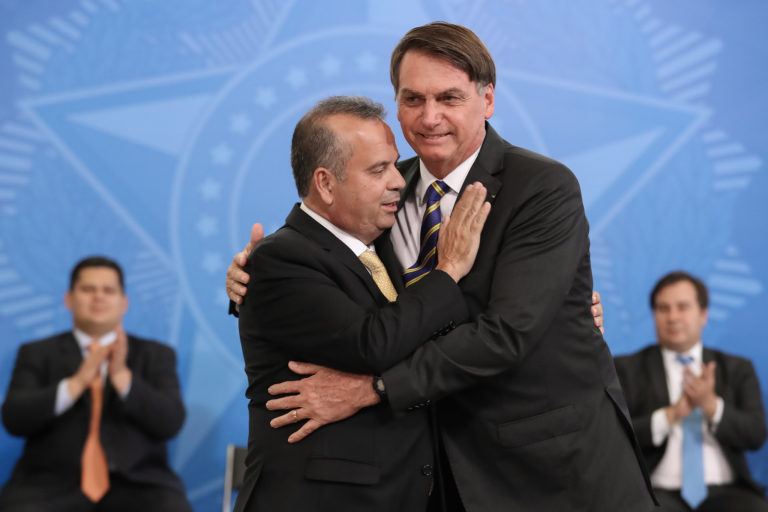 Rogério Marinho abraçando Jair Bolsonaro se abraçando, ambos com terno preto, camisa branca, com fundo azul e pessoas aplaudindo.