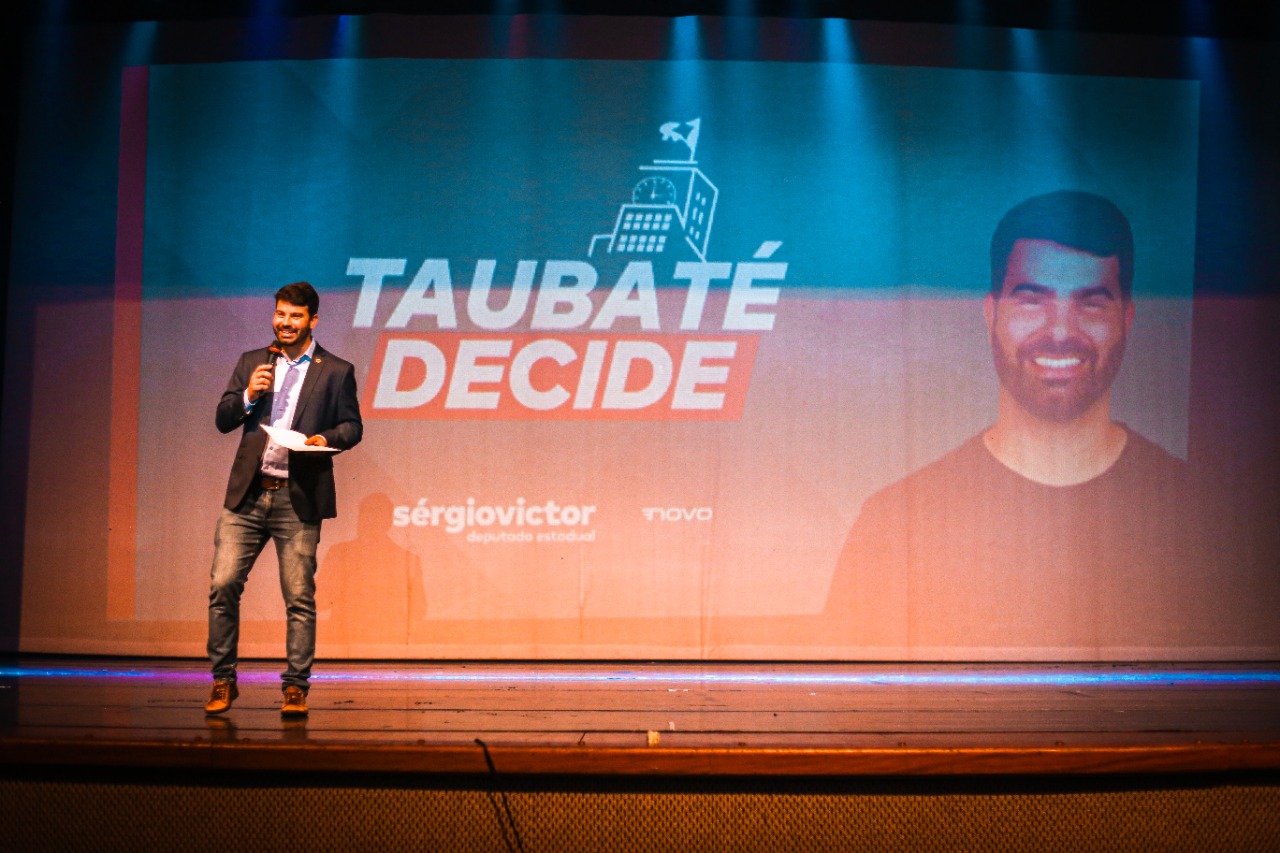 Deputado estadual Sérgio Victor em frente a um telão do Taubaté Decide, com roupa preta, em cima de um palco com luzes com detalhes em laranja.