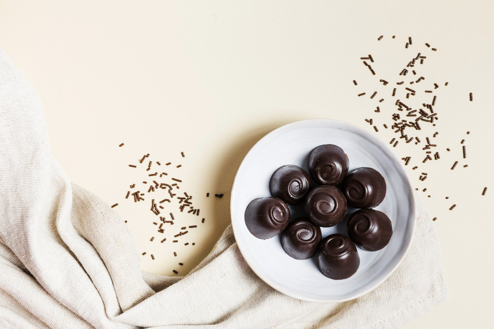 Bombons marrons em cima de um prato branco, com uma toalha e raspas ao lado, em cima de uma mesa branca.