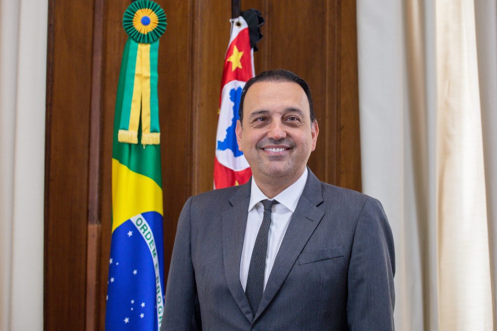 felicio ramuth, do tronco para cima, sorri de terno completo cinza com gravata cinza e camisa branca. ele sorri em frente às bandeiras do estado de são paulo e do brasil.