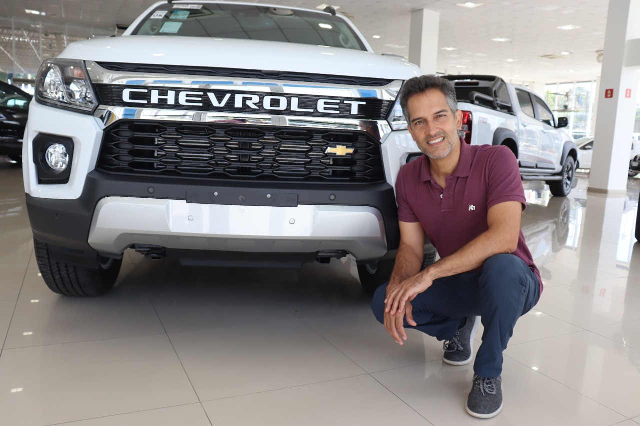 Rafael Davoli, diretor de Marketing do Grupo Davoli, em frente a um Chevrolet, com camisa polo roxa, calça jeans e sapato; foto ilustra matéria sobre inauguração da ACDelco em Caraguatatuba.