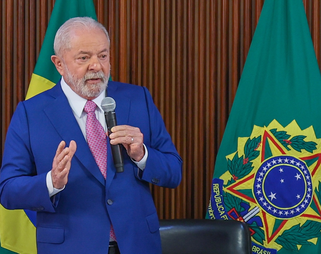 Presidente Lula com terno azul, camisa branca, gravata rosa, falando no microfone, em uma sala, com a bandeira do Brasil ao lado; foto ilustra matéria sobre o valor do auxílio-reclusão.