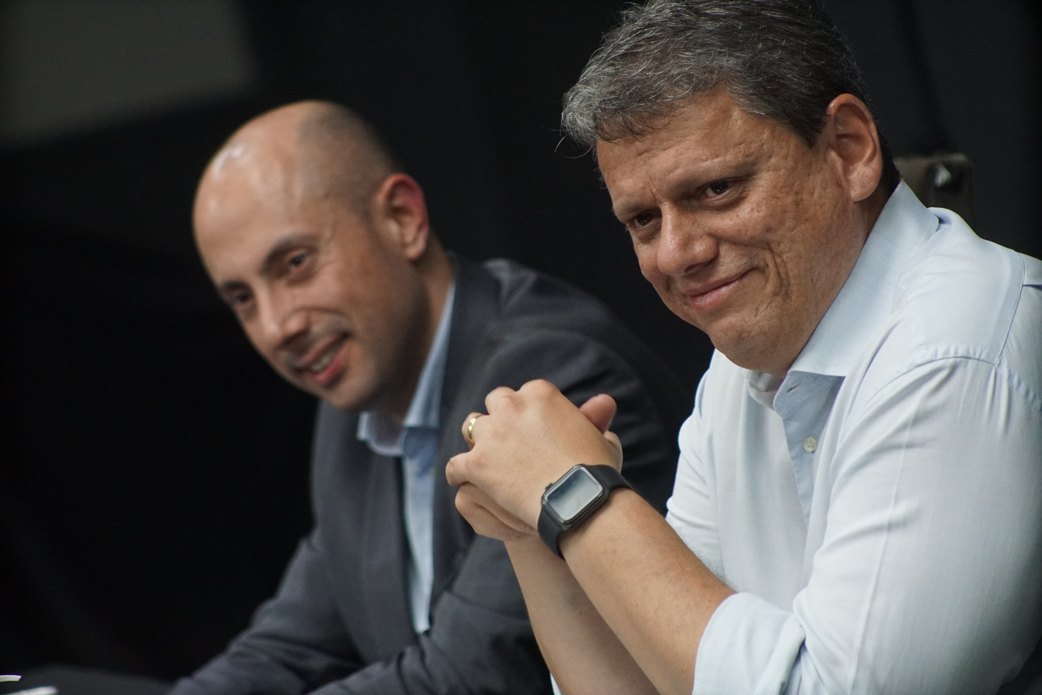 Tarcísio de Freitas durante reunião sobre IPVA com representantes do setor automotivo; na foto o governador de São Paulo usa camisa branca, relógio preto e olha para o lado, sorrindo; uma pessoa à sua esquerda usa terno cinza e camisa azul. O fundo é preto.