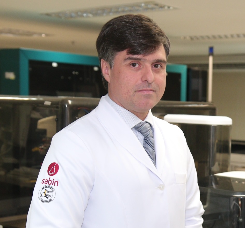 Dr. Alexandre Cunha, médico do Grupo Sabin, com jaleco branco e gravata cinza.