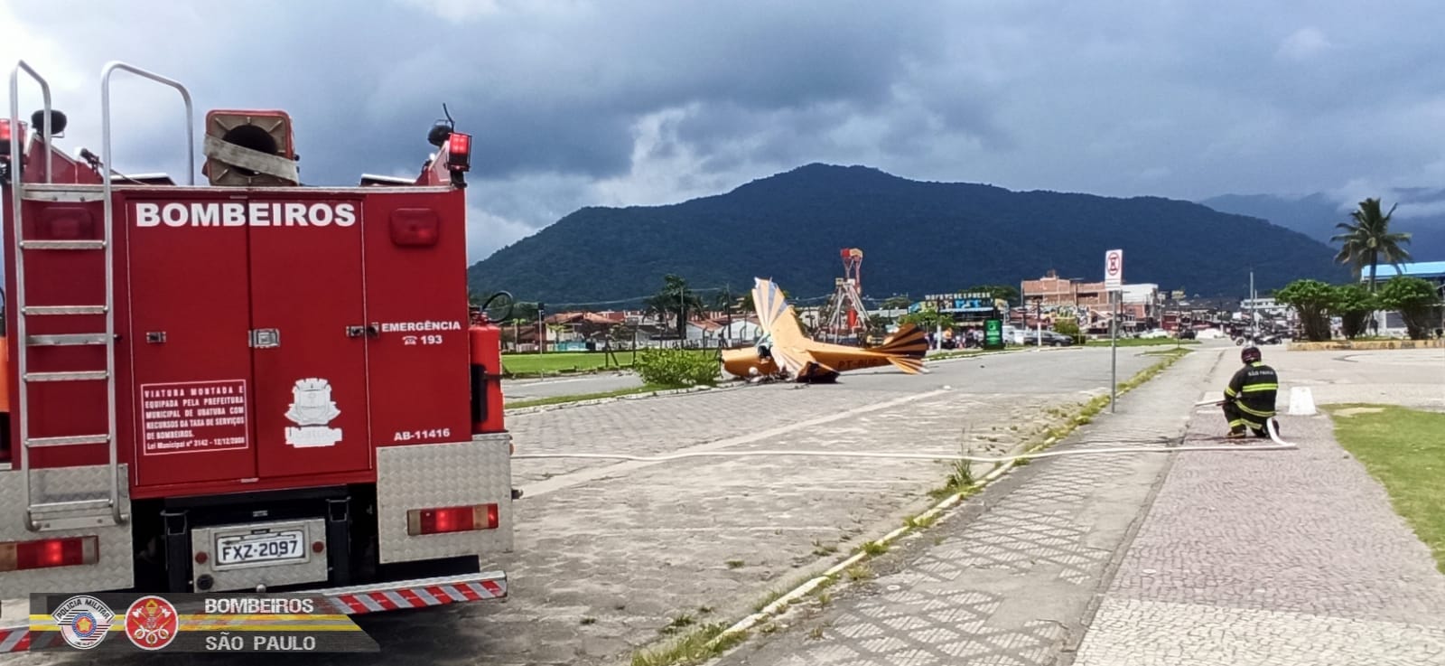 Foto mostra um avião de pequeno porte que caiu em Ubatuba, com céu azul, pista no meio, bombeiro ao lado e montanhas ao fundo.