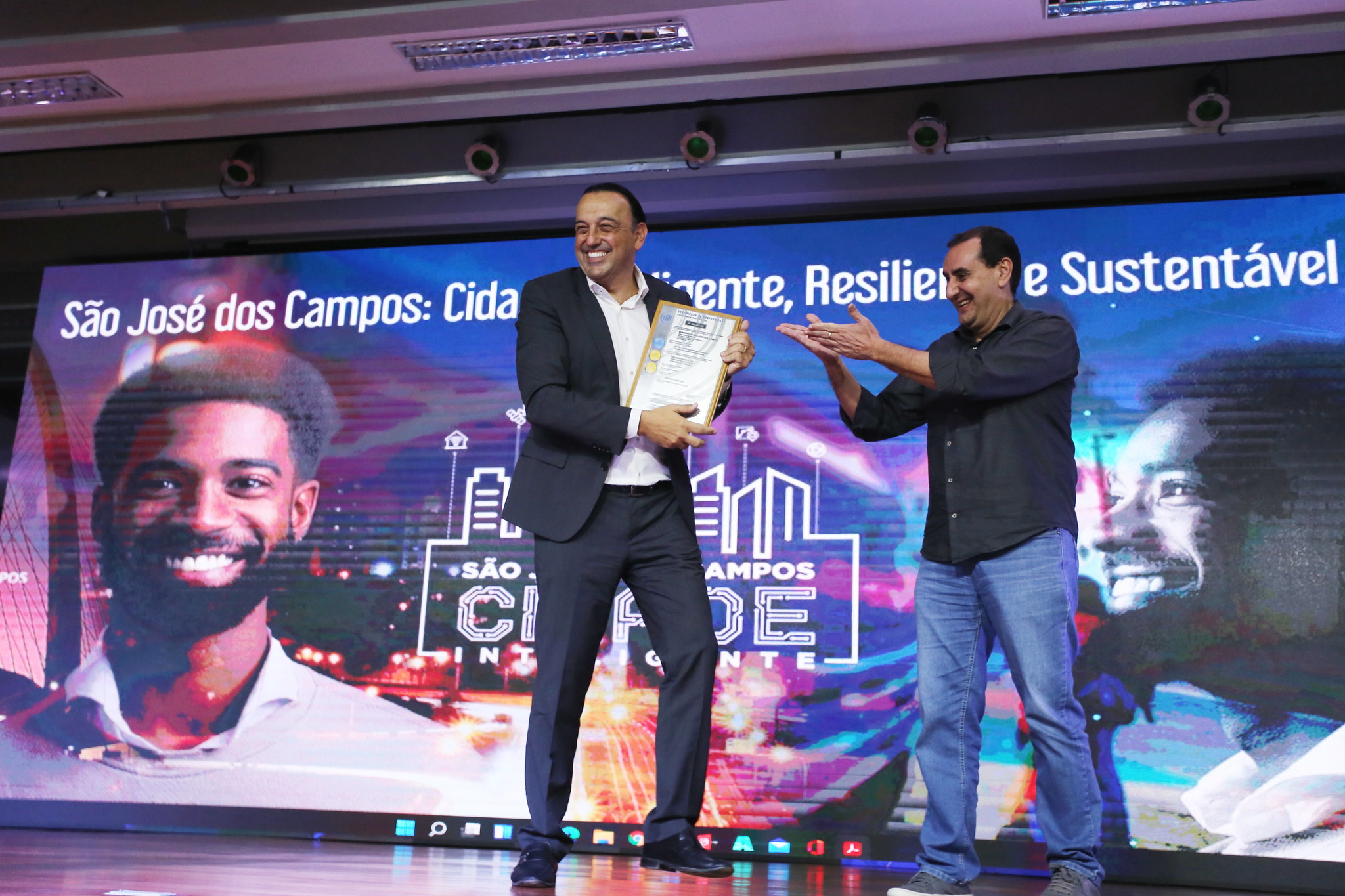 Felicio Ramuth segurando o certificado de Cidade Inteligente, que a ABNT deu para São José dos Campos (SJC), ao lado do seu então vice-prefeito, Anderson Farias, ambos do PSD. Foto mostra painel roxo, teto iluminado e os dois em cima do palco.