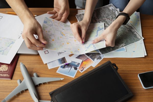 imagem de mãos de duas pessoas, planejando supostamente uma viajem, com um mapa. Tem também cartões postais e passaporte; foto ilustra matéria de cursos de qualificação sobre turismo, em Jacareí.