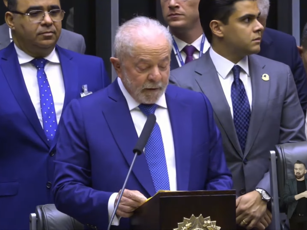 Lula durante discurso na cerimônia de posse, com terno preto, gravata azul, camisa branca, falando no microfone, com pessoas em volta.