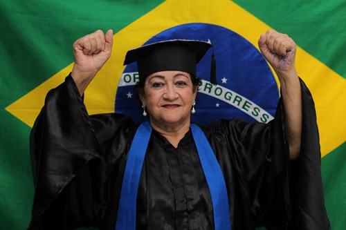 Aluna da EJA em São José dos Campos, com roupa de formatura azul e preta, em frente a uma bandeira do Brasil.