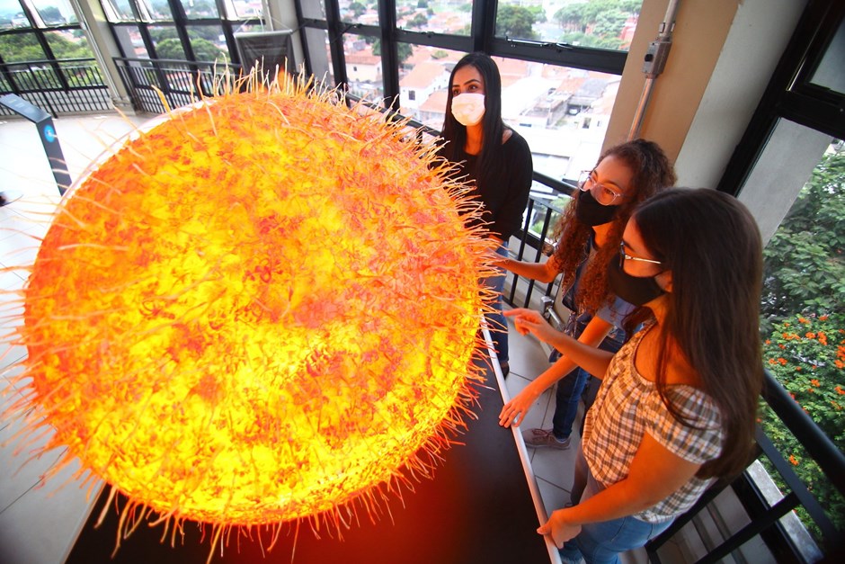 Pessoas dentro do Museu Interativo de Ciências, em SJC, em volta de um objeto laranja; foto ilustra matéria sobre Férias no MIC.