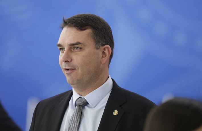 Flávio Bolsonaro com terno preto, gravata preta, camisa branca, em um fundo azul e olhando para o lado; senador votou contra a intervenção federal em Brasília.
