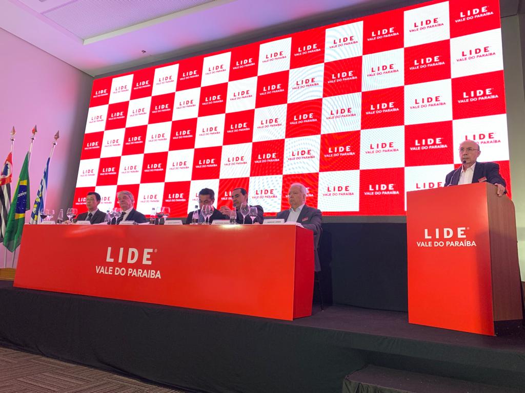 Henrique Meirelles discursando durante o evento do Lide Vale do Paraíba, no Colinas Shopping, em São José dos Campos; foto tem painel vermelho ao fundo e pessoas sentadas em volta.