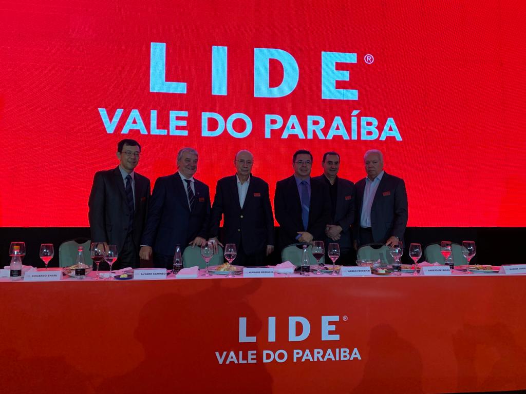 Empresários do Lide Vale do Paraíba participam de encontro com Henrique Meirelles, em SJC, com painel vermelho ao fundo e a marca da empresa na frente.