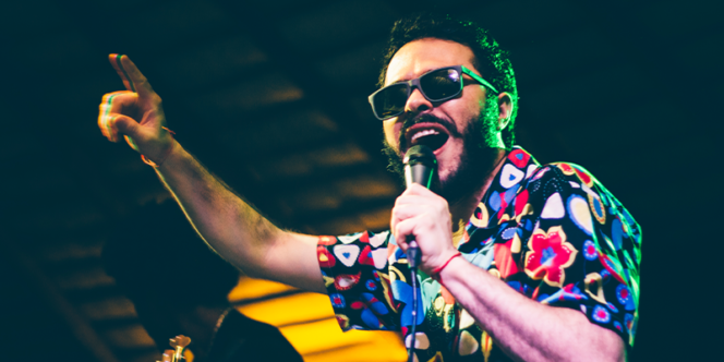 Jottape & Quarteto São Jorge, que tocará no SESC SJC. Foto mostra o cantor com camisa florida, falando no microfone, óculos escuros e braços levantados.