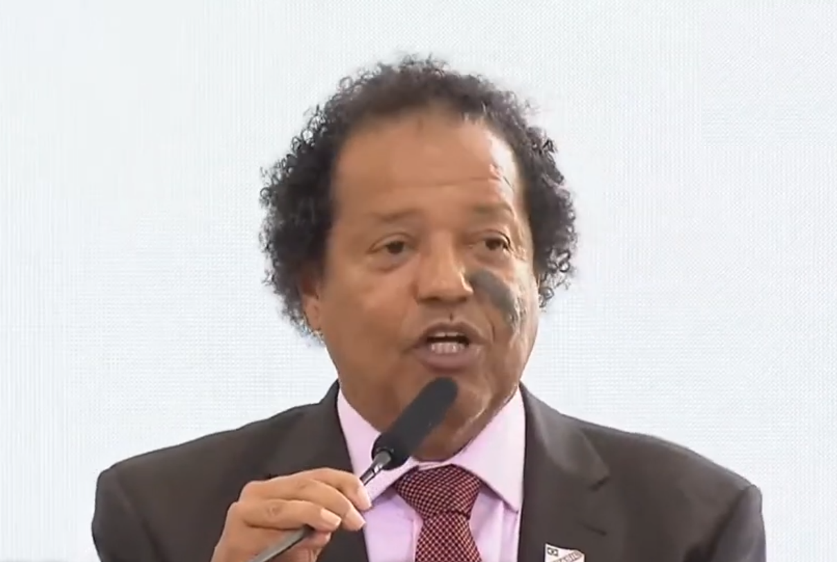 Luiz Carlos Mancha com terno marrom, camisa rosa e gravata vermelha, falando no microfone durante evento com Lula; foto ilustra matéria em que ele defende o socialismo.