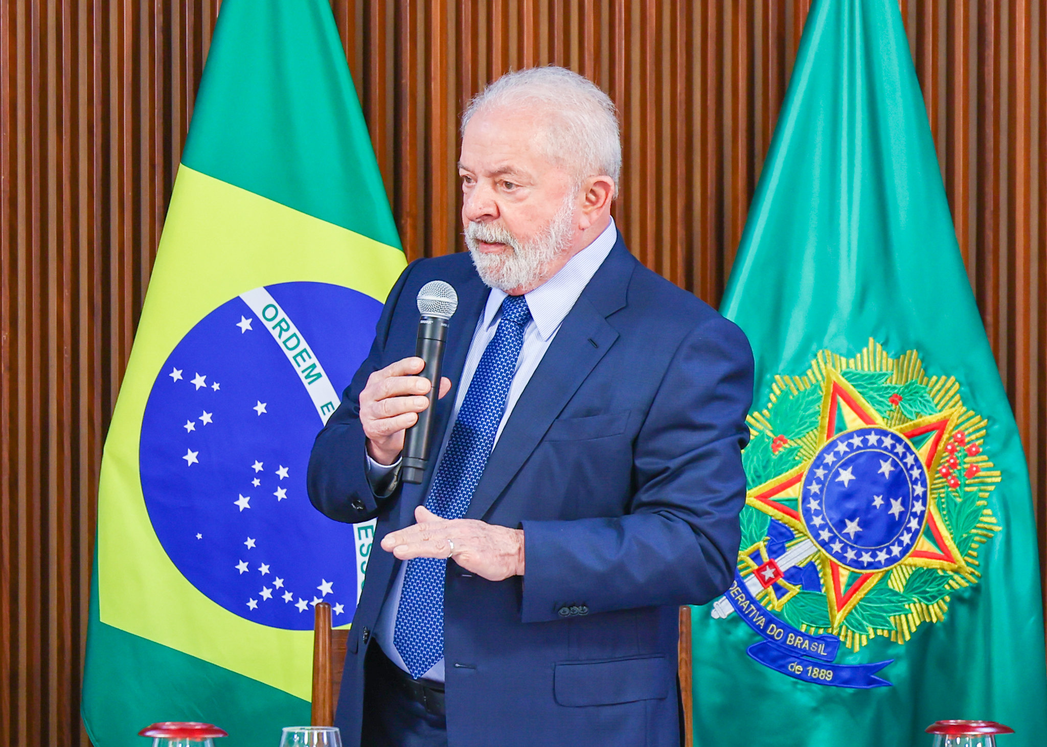 Presidente Lula durante reunião com governadores, usando terno preto, gravata azul, camisa branca, em frente a uma parede marrom e bandeiras do Brasil.