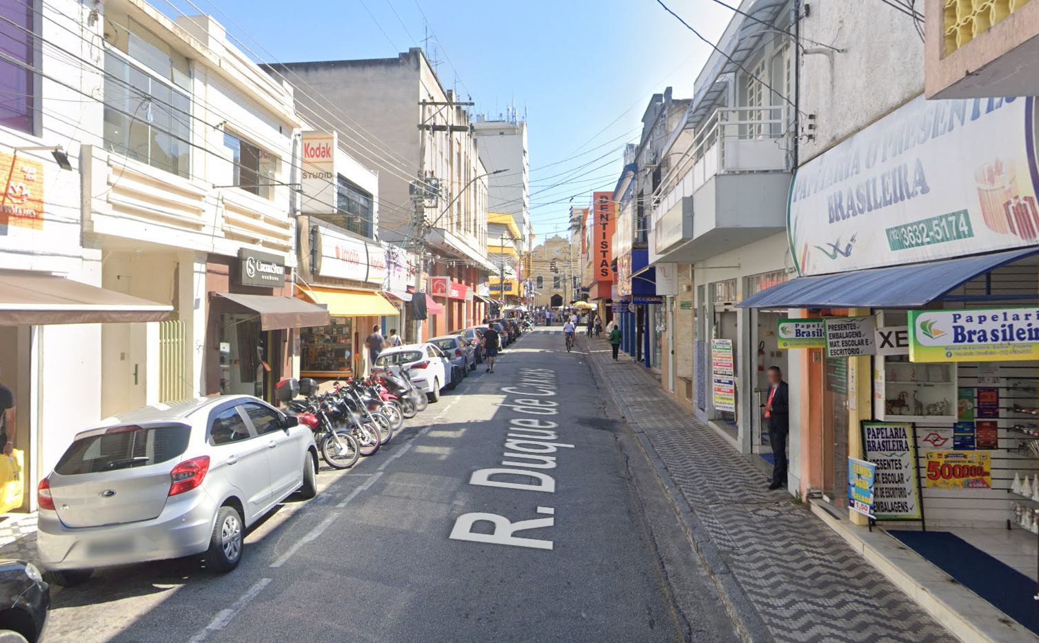 Rua Duque de Caxias, no Centro de Taubaté, com carros do lado, lojas e uma igreja ao fundo.