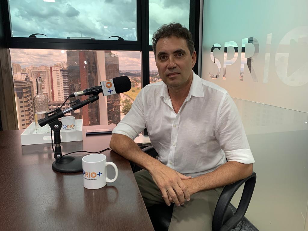 Sergio Theodoro, diretor-executivo da Agemvale, durante entrevista sobre as câmeras de segurança, o Cinturão Eltrônico. Na foto ele está sentado, falando no microfone, com uma mesa, uma caneca e a imagem de SJC ao fundo.