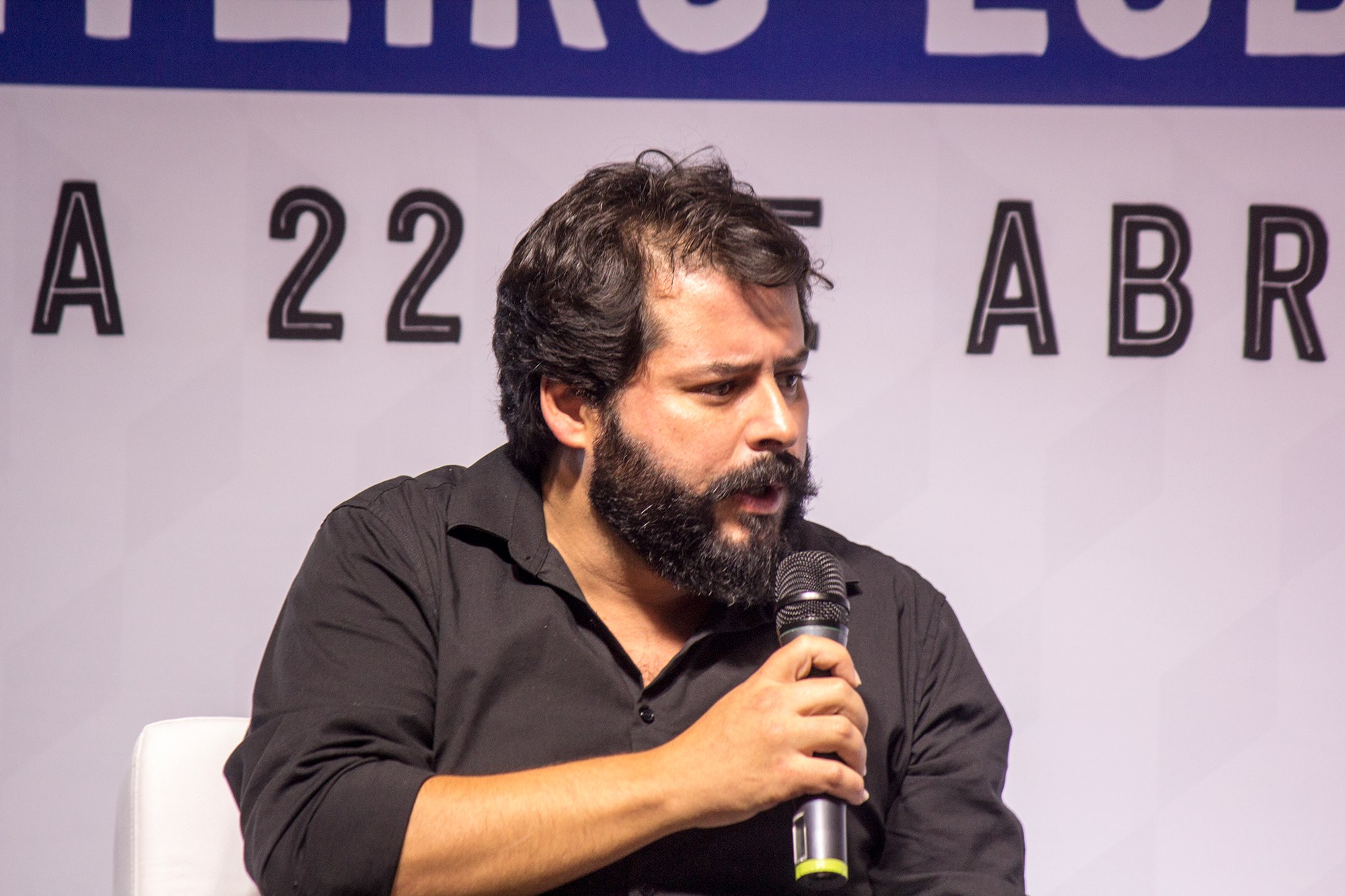 Escritor Tiago Feijó, de Guaratinguetá, fala no microfone, com fundo branco, camisa preta, barba e olhando para o lado.