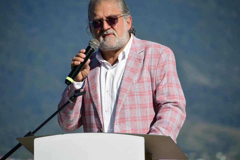 Toninho Colucci, prefeito de Ilhabela, com terno rosa, camisa branca, falando no microfone, óculos escuro e fundo azul. Foto ilustra matéria sobre controle de entrada de turistas na ilha.