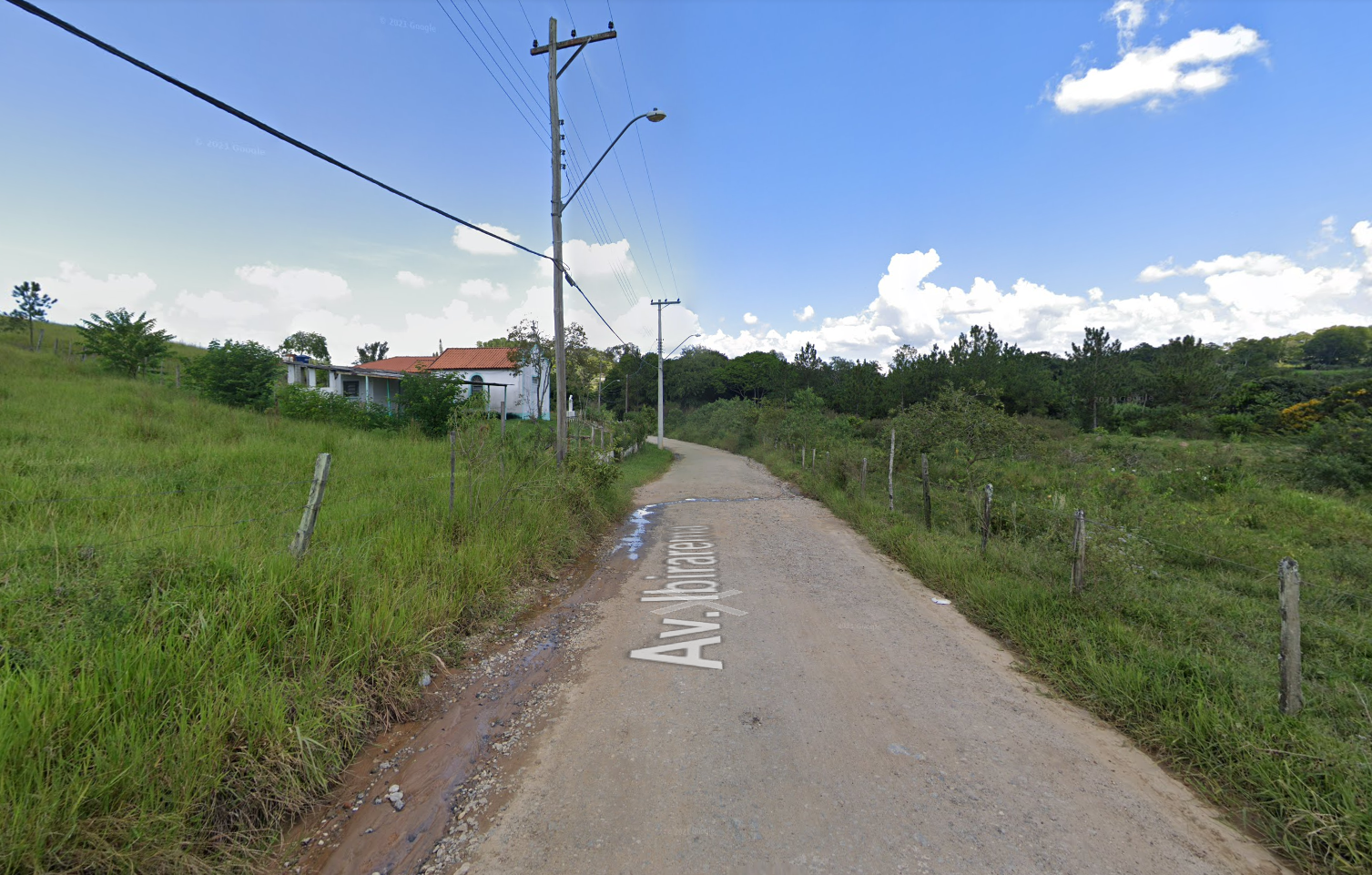 Bairro Veraneio Irajá, zona rural de Jacareí, com céu azul, área verde nas laterais, estrada de barro, poste e uma casa ao fundo.