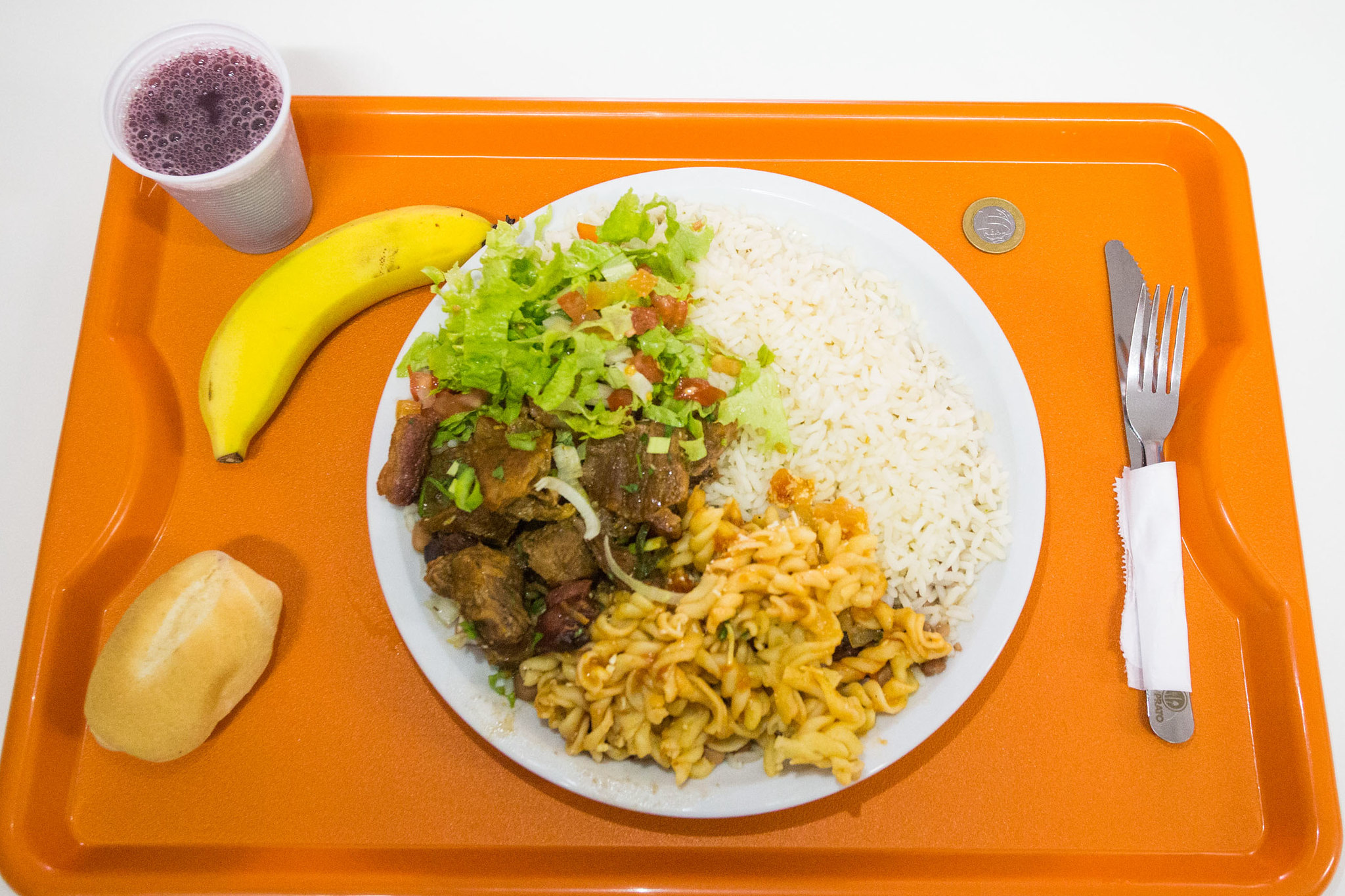 foto tirada de cima de prato do bom prato. refeição tem salada, arroz, carne, macarrão, e bandeja incluiu ainda talheres, pão, banana e suco.