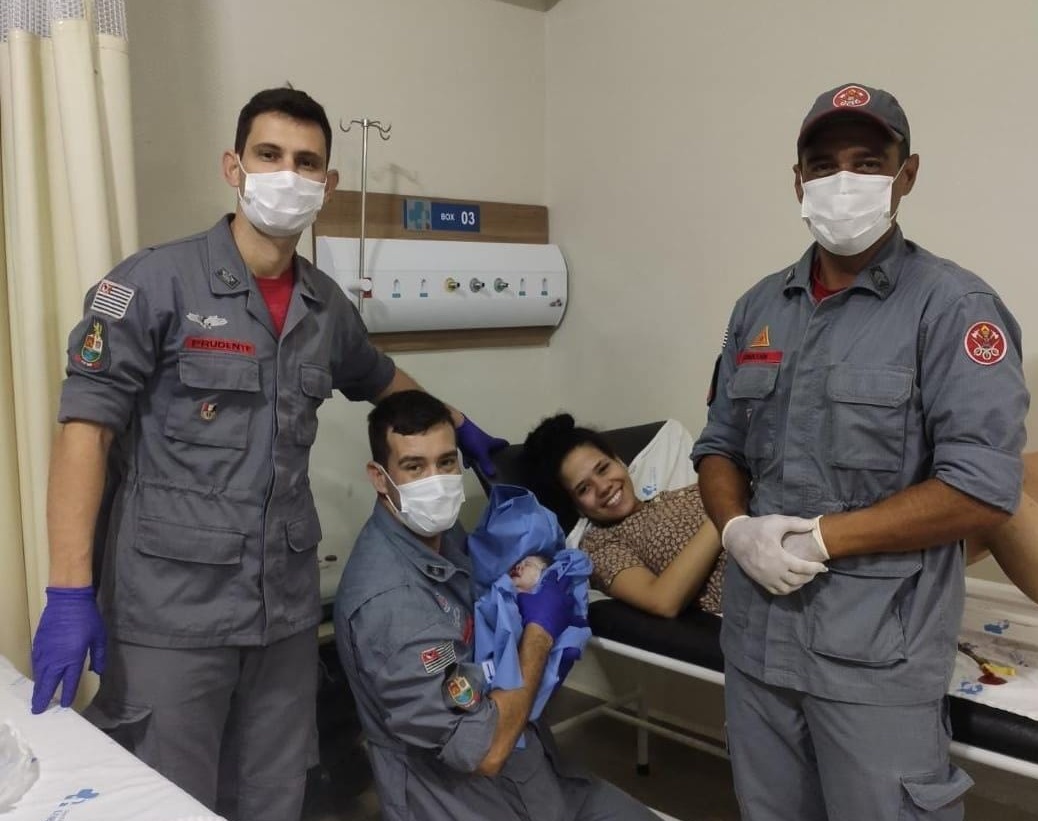 Foto mostra bombeiros de Lorena auxiliando em parto de mulher na cama. Os homens usam roupas cinzas, máscara e luvas; enquanto a mulher está deitada; já o bebê usa roupas azuis.