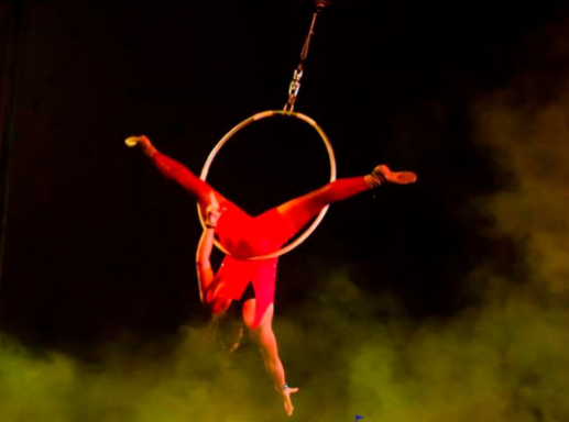 Holliday Circus é opção de Circo em Taubaté; foto mostra mulher com roupa vermelha, fundo preto, fumaça amarela e pendurada em um acessório circense.