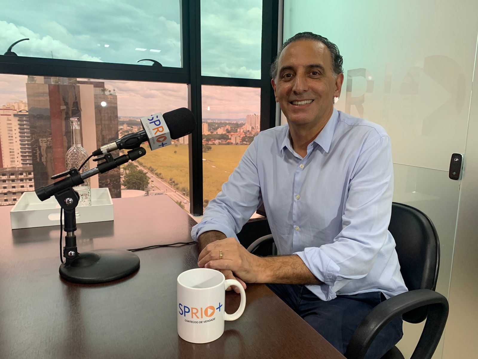 Retrato do Eduardo Cury sentado, com uma camisa azul, sorrindo, falando no microfone e a paisagem de São José dos Campos ao fundo.