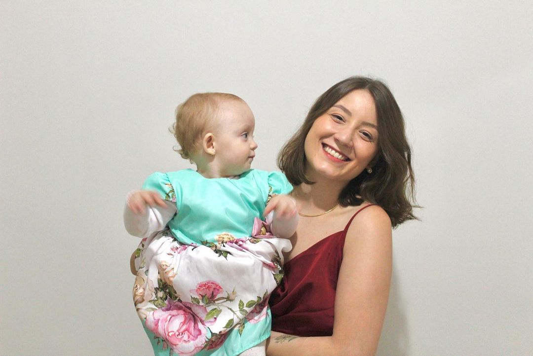 Isabelle Mioni e sua filha; foto ilustra matéria sobre jovem de SJC que acusou o Hospital da Vila Industrial, gerido pelo SUS, de violência obstétrica. Na foto ela veste blusa vermelha e segura sua filha no colo, enquanto sorri para a câmera, já a filha usa vestimentas verdes e olha para o lado.