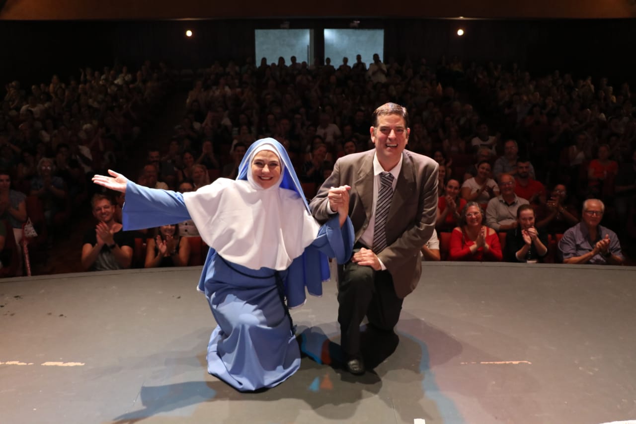 Peça espírita “Divaldo e Joanna”, que chega ao Teatro Colinas neste fim de semana. Foto mostra mulher com traje azul e branco, homem de terno marrom ao seu lado e plateia ao fundo.