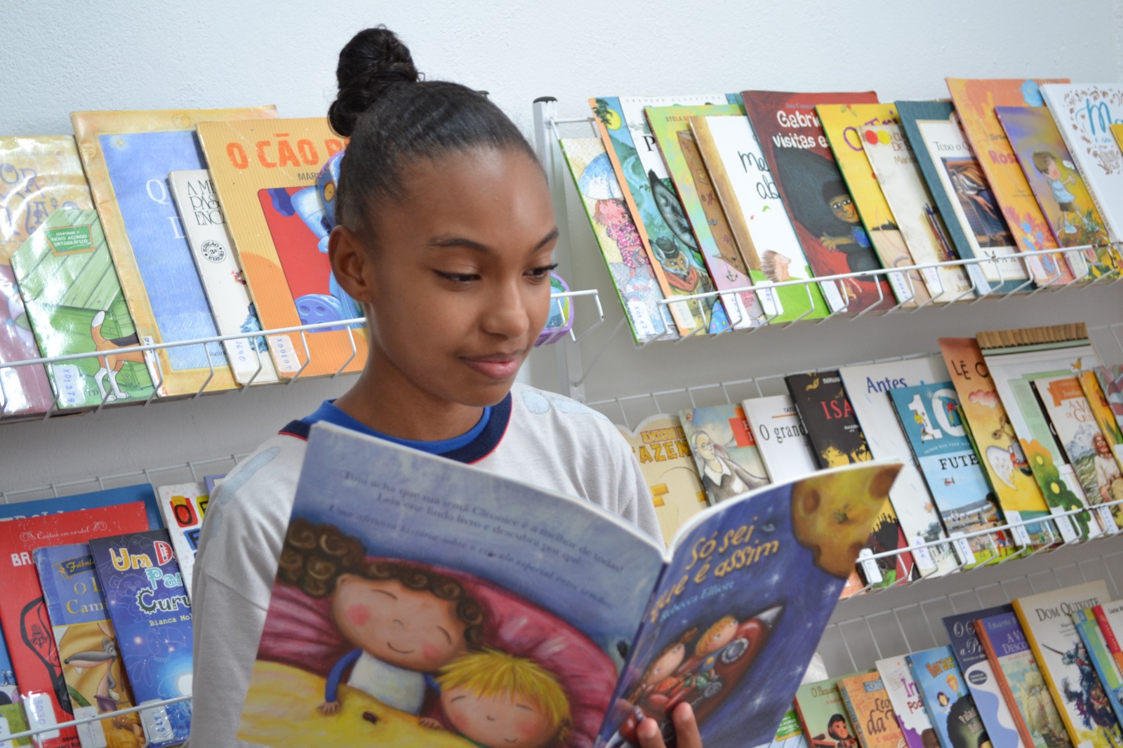 Projeto Portas Abertas Para o Saber, em São José dos Campos, atende crianças com deficiência; foto mostra garota lendo um livro azul, vestindo camiseta branca, em frente a uma estante com outros livros.
