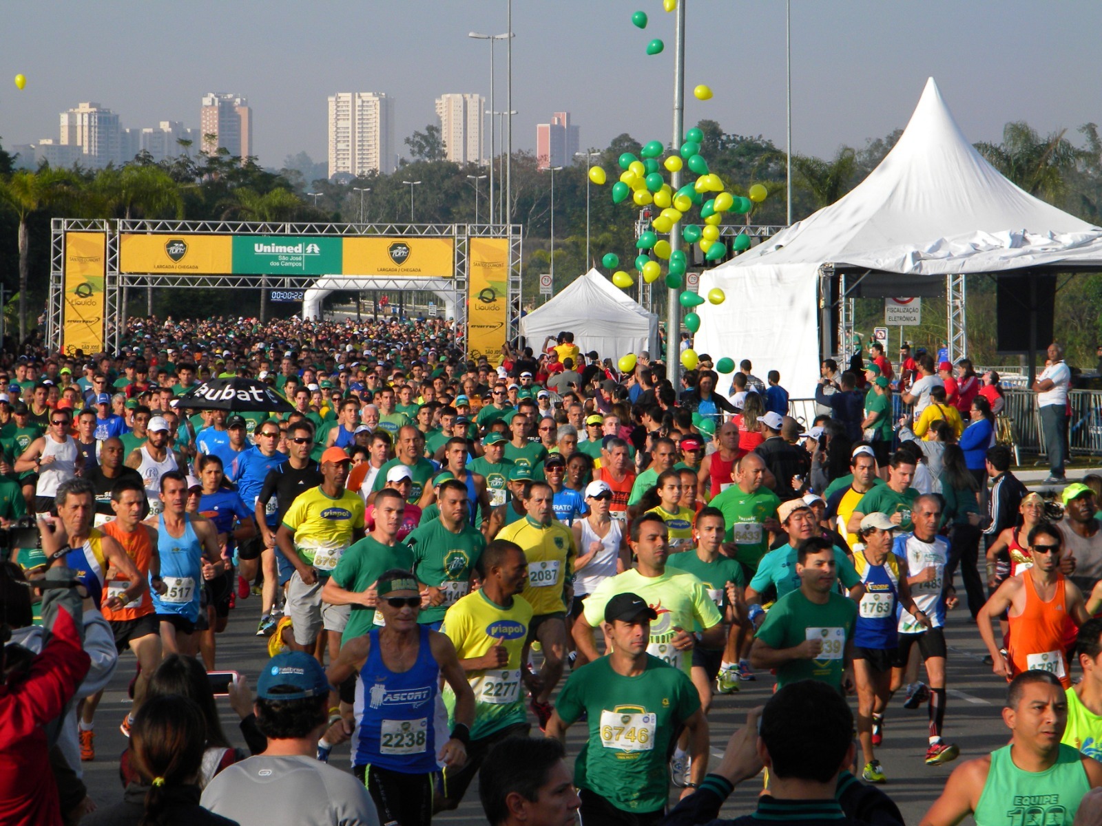 Unimed Run 2023 acontecerá em São José dos Campos em março; foto mostra pessoas com camisetas verdes, amarelas e azuis, com céu azul, tenda branca na direita, balões voando e um painel ao fundo.