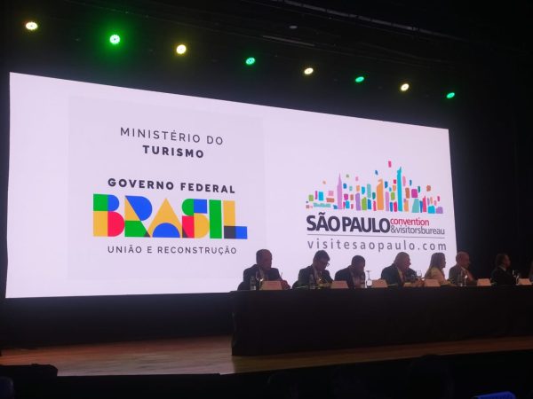 evento de turismo em sp. foto mostra palco com mesa e telão com logomarca do governo federal.